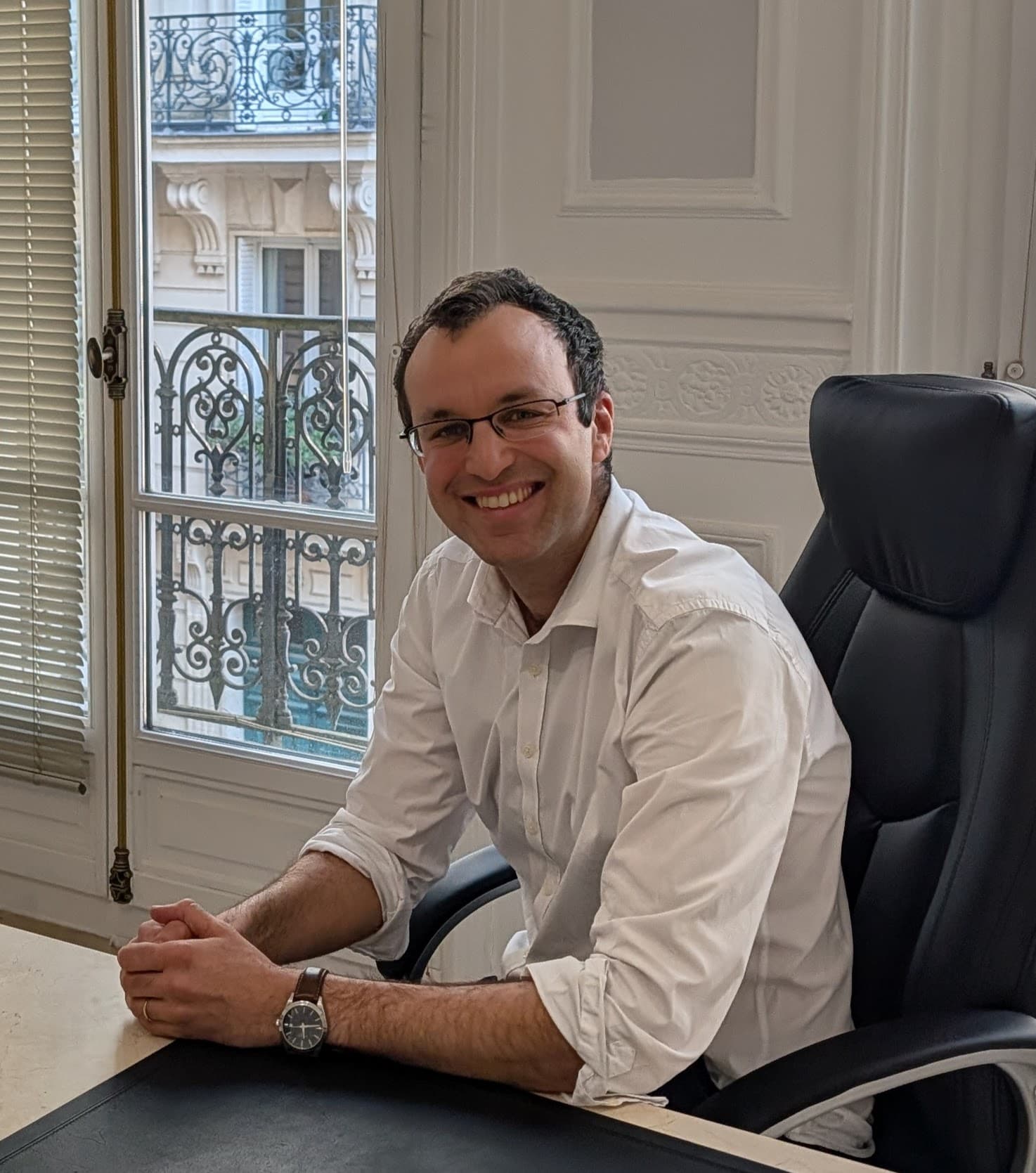 Homme souriant à lunettes et chemise blanche assis à son bureau devant une fenêtre.