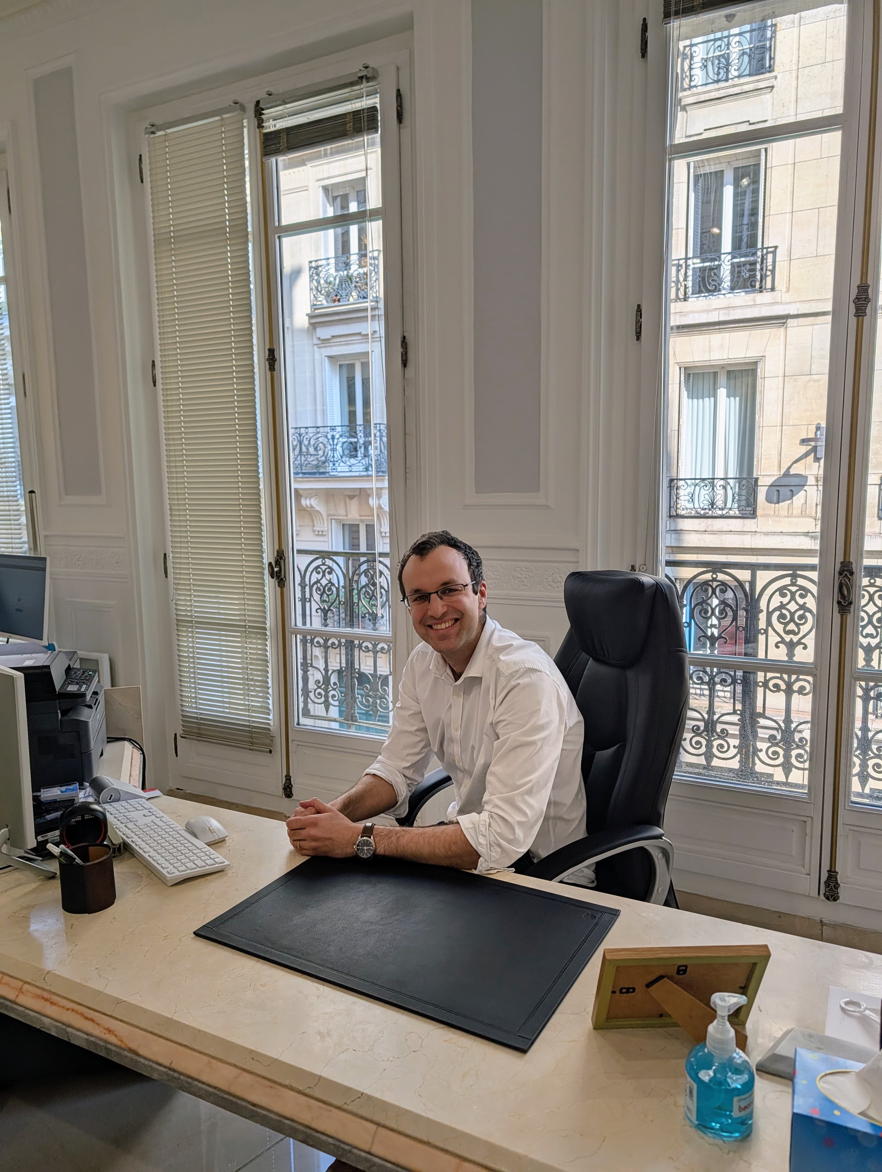 Homme souriant en chemise blanche assis à son bureau devant de grandes fenêtres de ville.