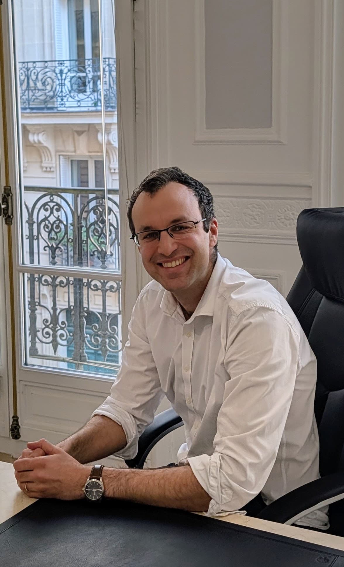 Homme souriant à lunettes et chemise blanche assis à son bureau devant une fenêtre.