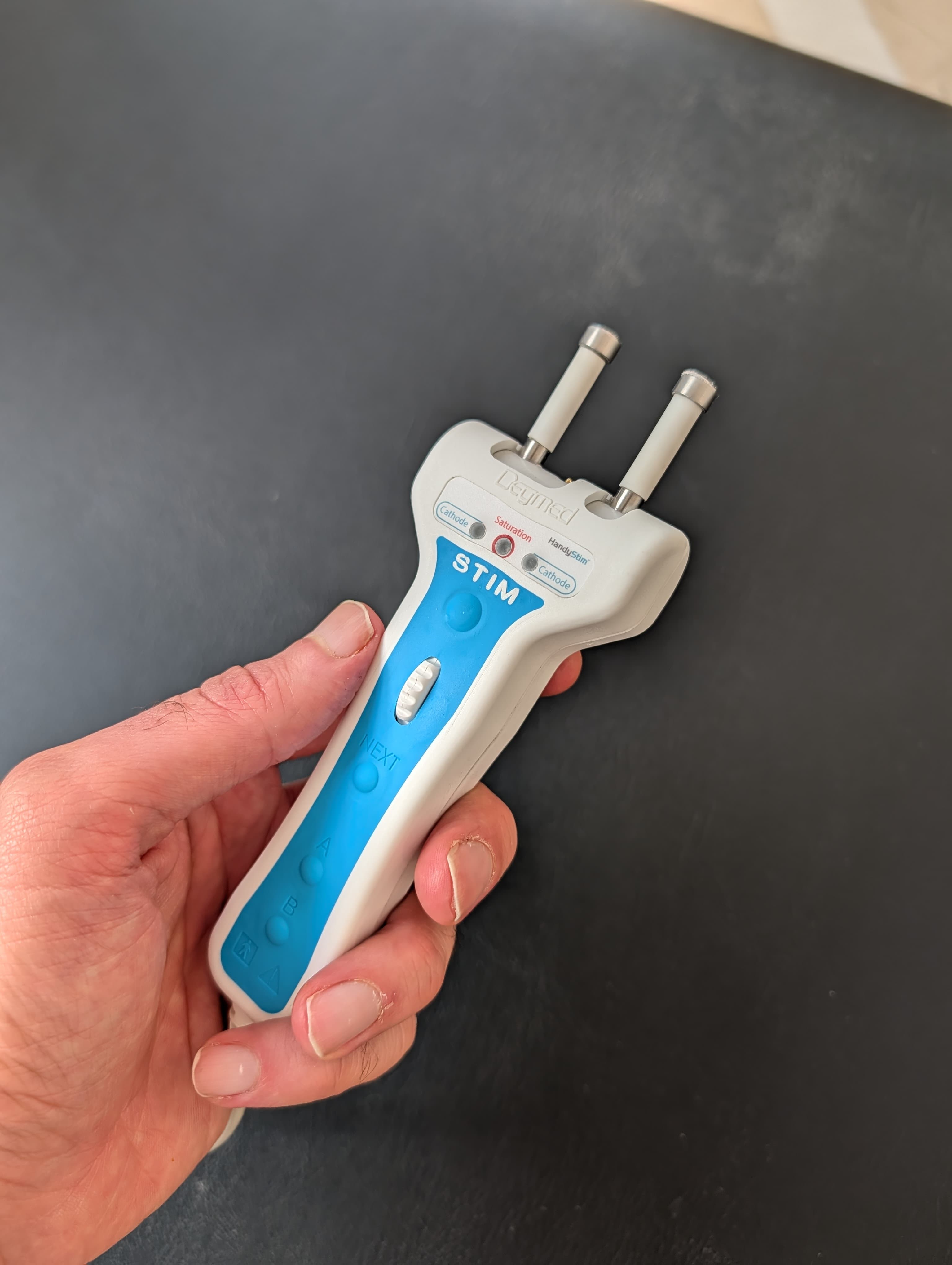 Main tenant un stimulateur nerveux Deymed blanc et bleu avec deux électrodes sur fond noir.