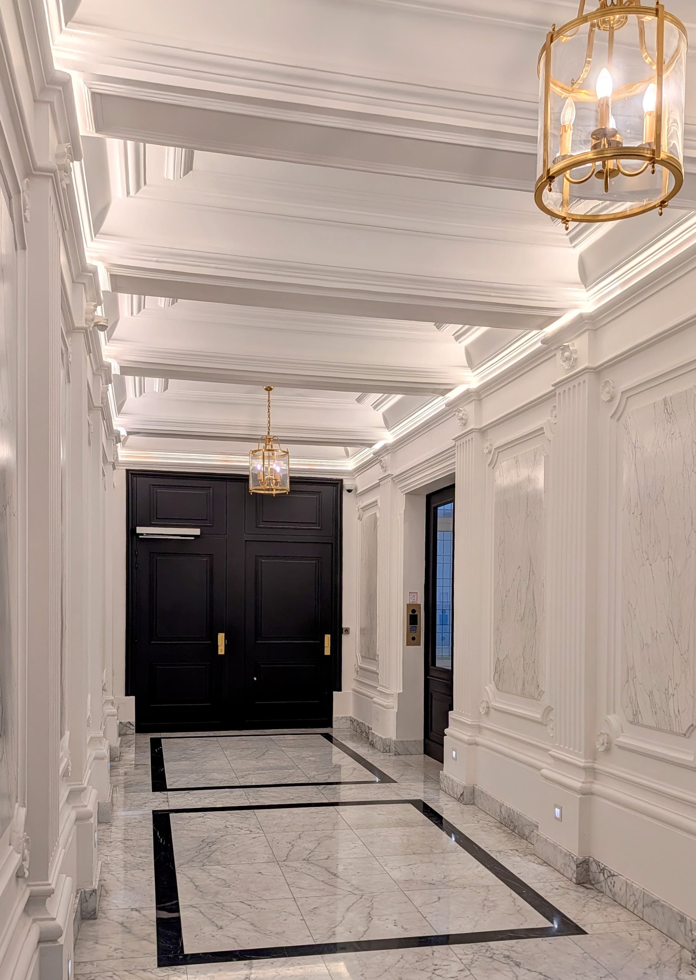 Couloir luxueux aux murs blancs moulurés, lustres dorés et grandes portes noires au fond.