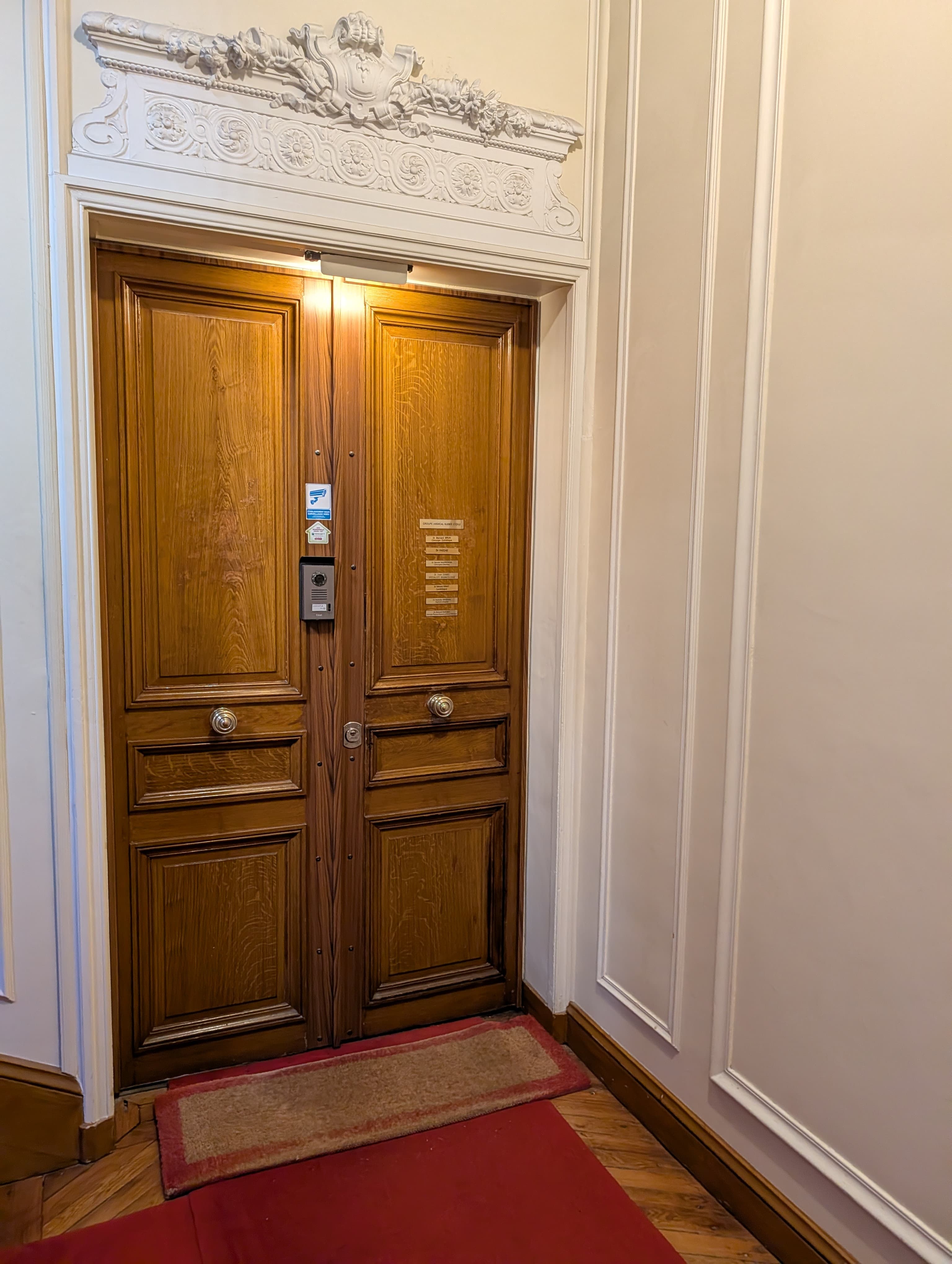 Portes doubles en bois avec moulures blanches sculptées, un interphone et tapis rouge.
