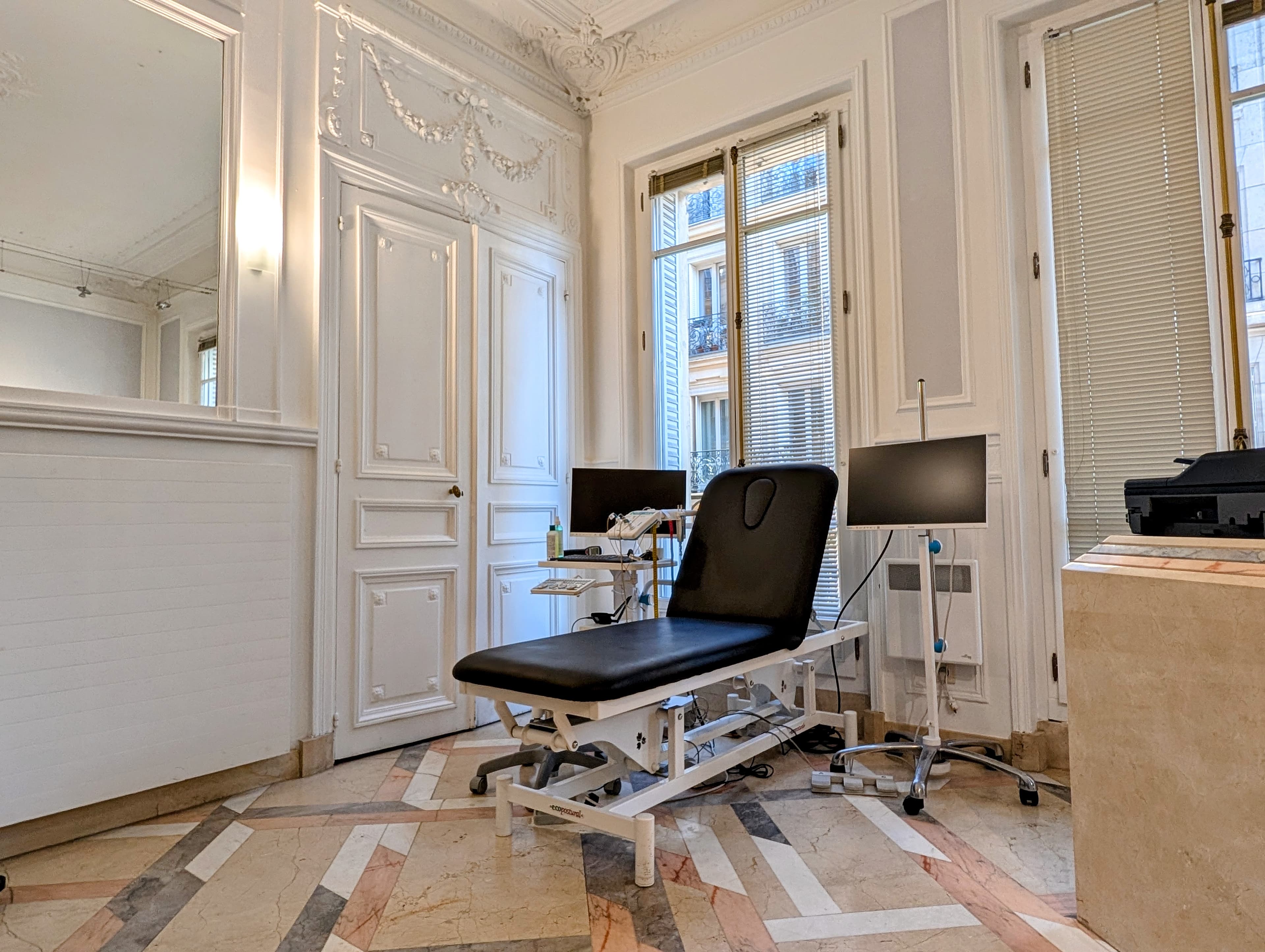 Table d'examen noire dans un cabinet médical élégant aux murs blancs ornés de moulures.